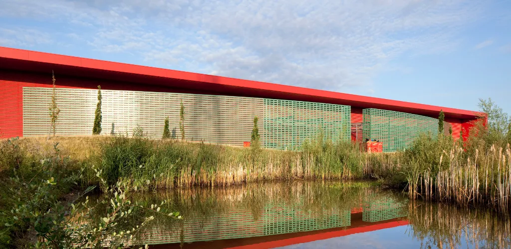 Bâtiment à toiture rouge et façade ajourée verte, se reflétant dans un bassin bordé de roseaux